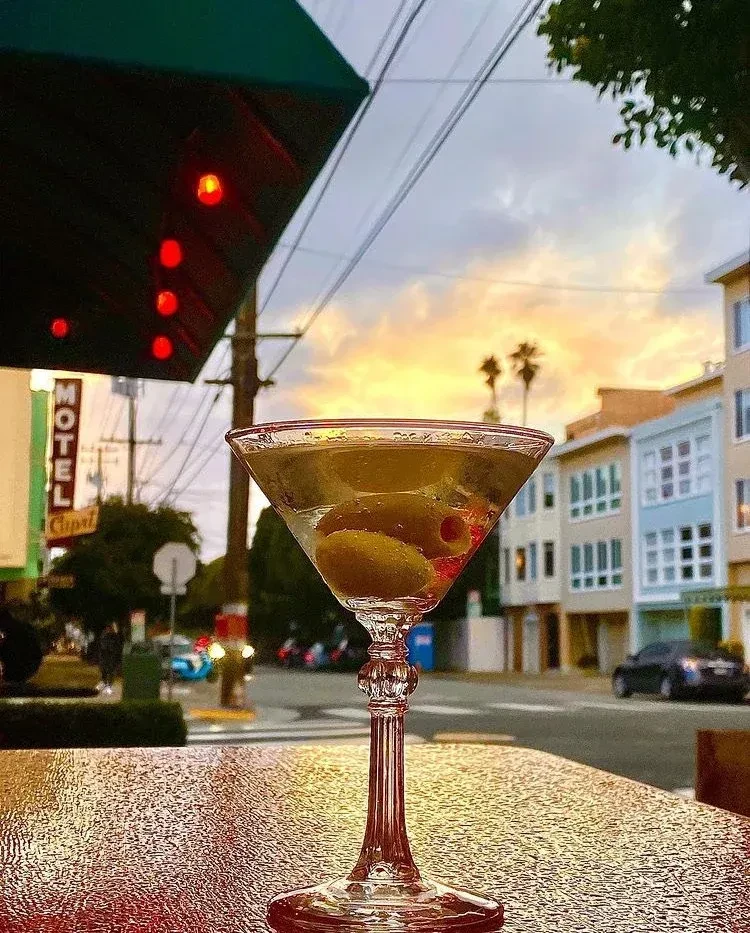 A martini glass with olives sits on a table outdoors at sunset.