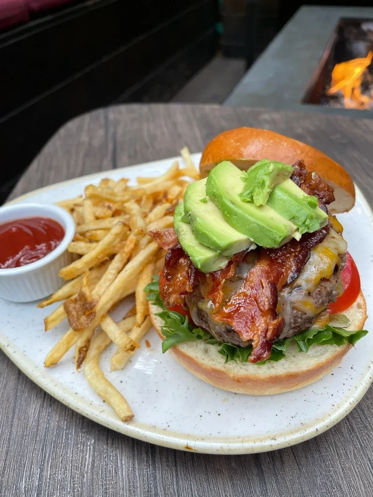 Close-up of gourmet burger with avocado, bacon, fries, ketchup on wood.