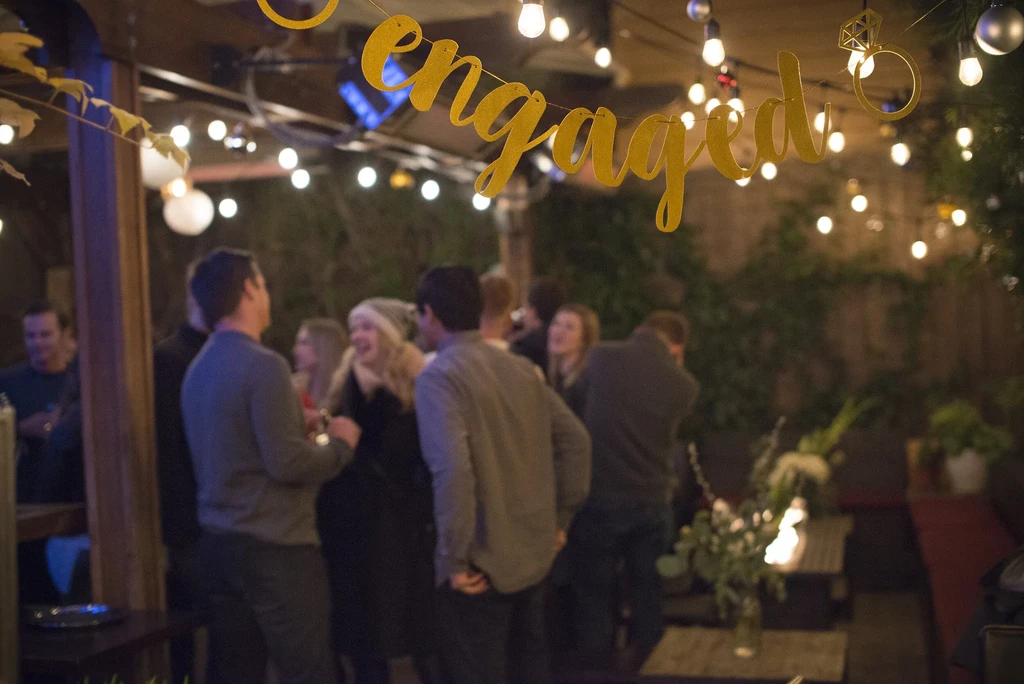 Outdoor engagement party with blurred people, string lights, and 'engaged' banner.