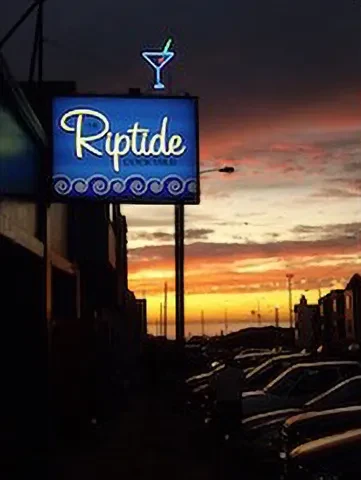 A neon "Riptide" sign with a martini glass against a vibrant sunset.
