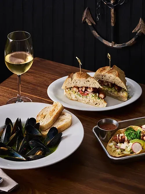 Mussels, sandwiches, tacos, and a glass of white wine on a wooden table.