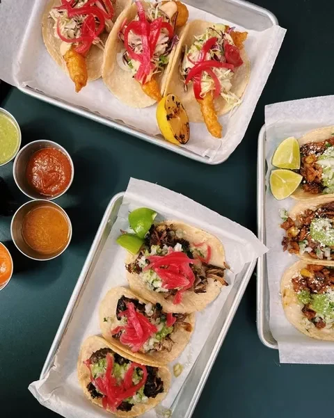 Three trays of tacos and three small cups of sauce on a dark table.