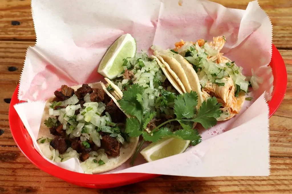 Three street tacos in a red basket with lime and cilantro.