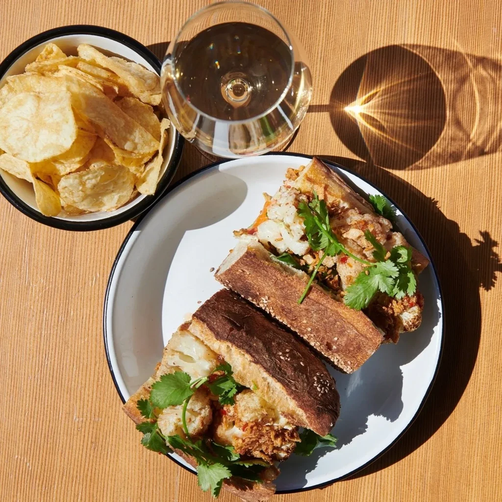 A crispy baguette sandwich with cilantro, chips, and a glass of white wine.