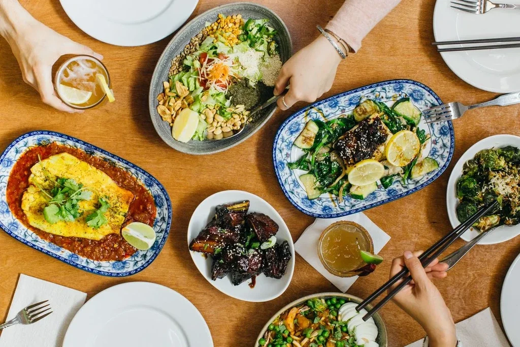 Overhead shot of many dishes, hands, and drinks on a wooden table.