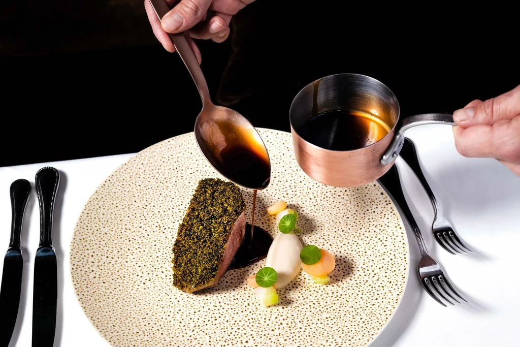 A chef pours sauce from a spoon onto a plated gourmet meal.