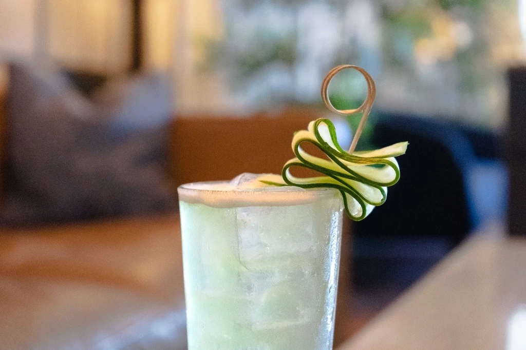 A light green cocktail with ice and a cucumber ribbon garnish.