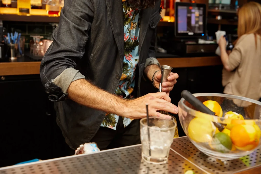 A bartender measures a drink while a woman works in the background.