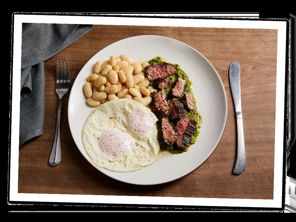 A white plate with steak, beans, and two fried eggs.