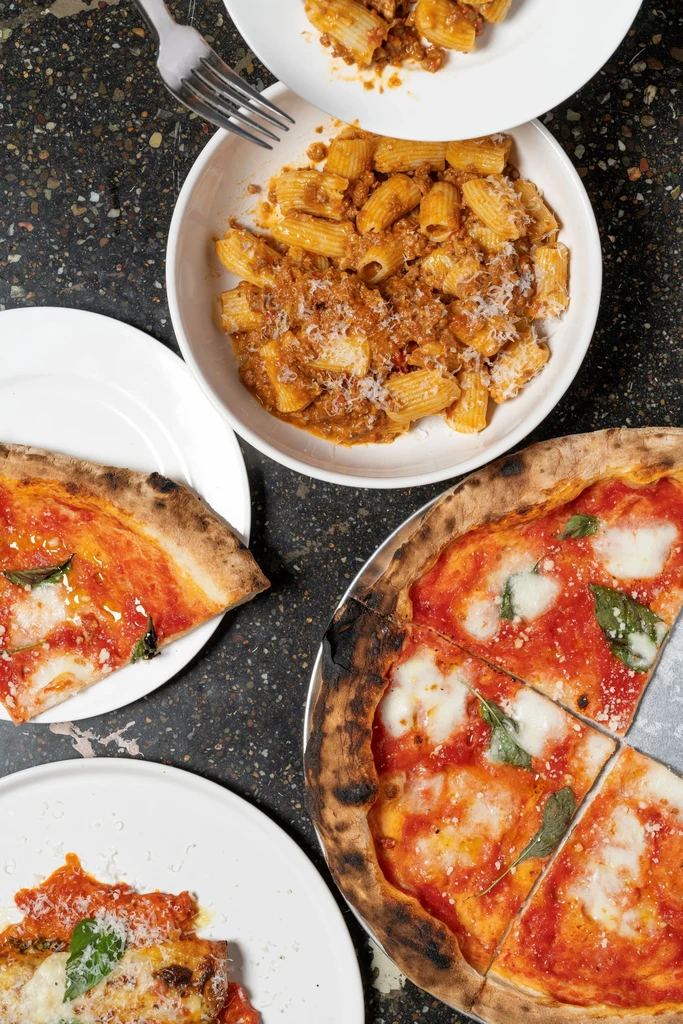 Overhead view of two pasta dishes and two pizzas on a speckled dark counter.