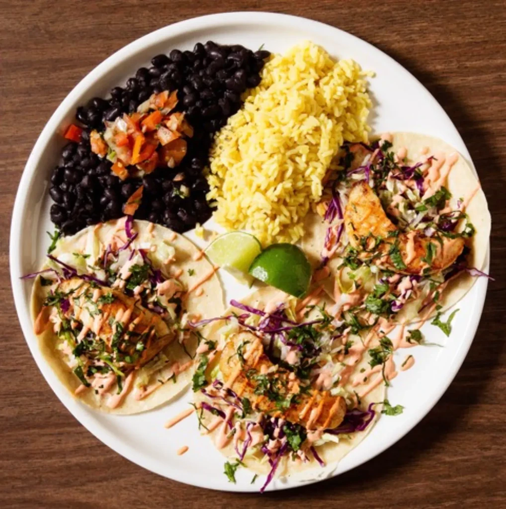 A white plate with three tacos, yellow rice, black beans, and pico de gallo.