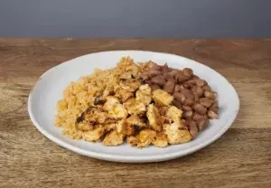 A plate of seasoned chicken, rice, and pinto beans on a wooden table.