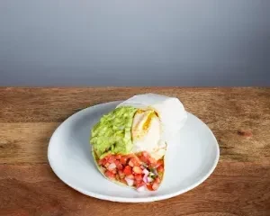 A white plate holds a half-wrapped burrito on a wooden table.