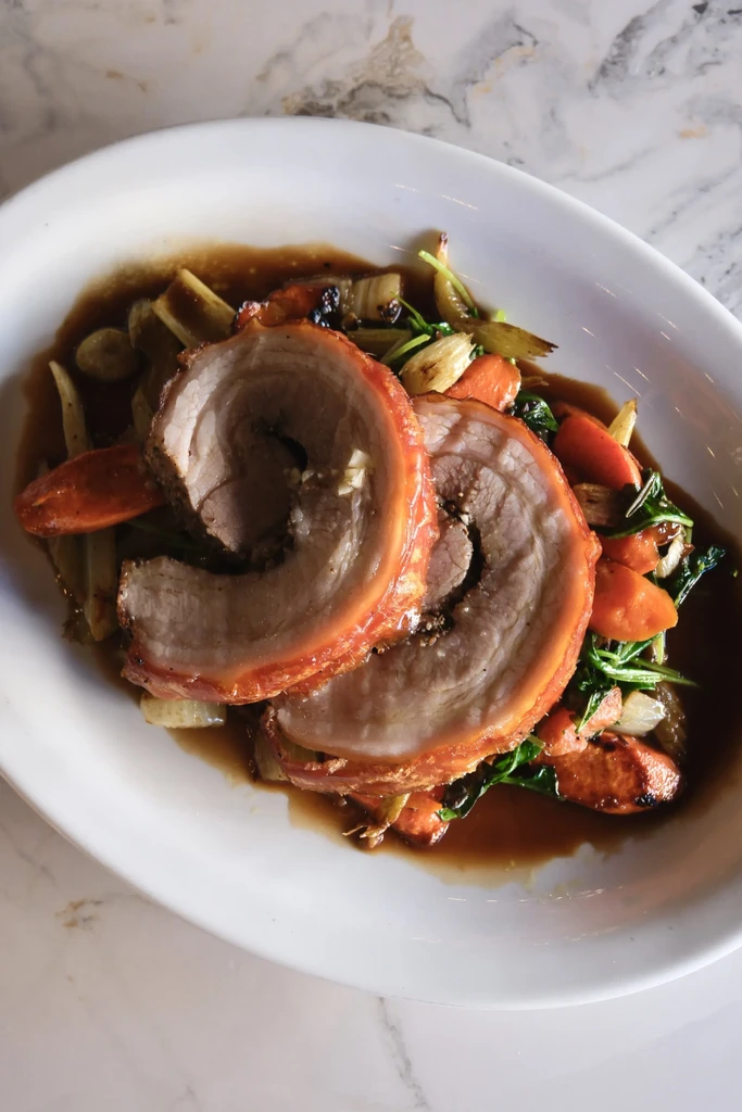 Two slices of rolled roasted pork belly, vegetables, and gravy on a plate.