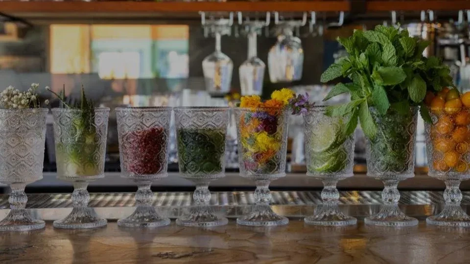 Eight ornate glasses filled with various ingredients on a bar counter.