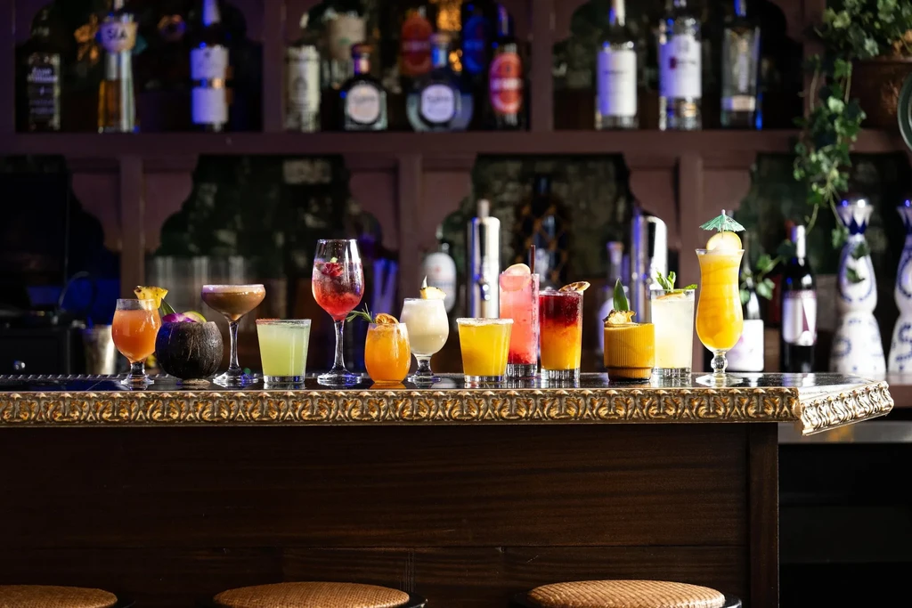 A bar counter with 12 colorful cocktails, liquor bottles in the background.