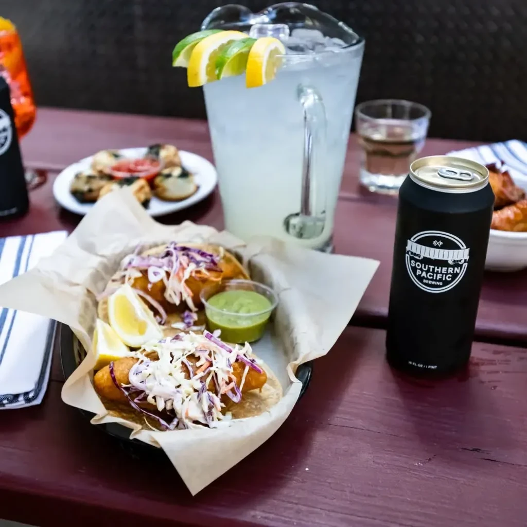 Fried fish tacos, a pitcher of lemonade, and a beer can on a table.
