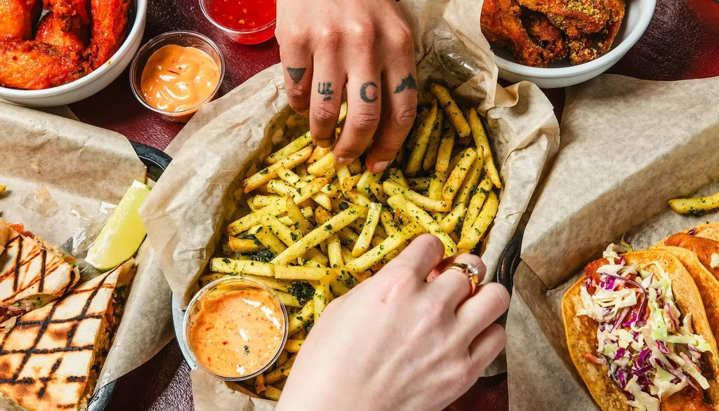 Two hands reach for fries in a basket with other fast food items.