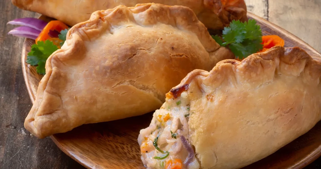 A wooden plate holds several golden brown savory pastries, one cut open.