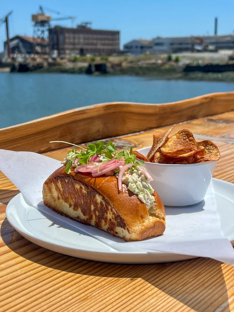 Lobster roll with pickled onions and chips next to water and industrial buildings.