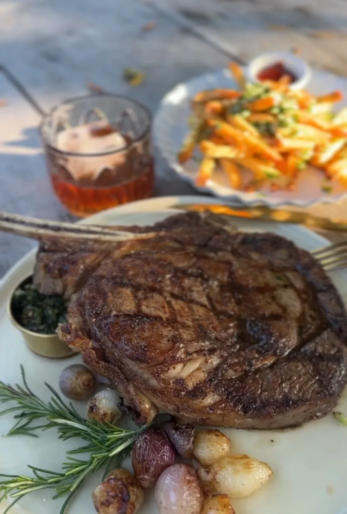 A grilled bone-in steak, fries, and a drink on an outdoor table.