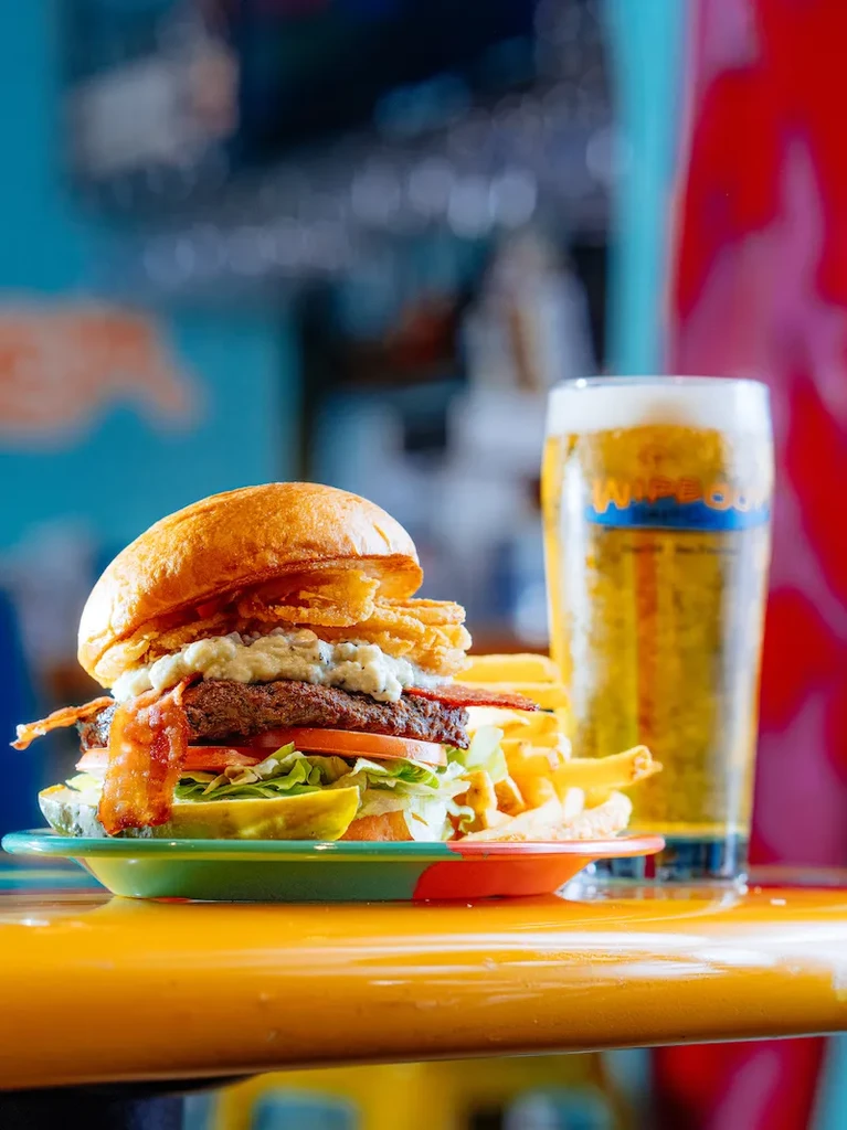A bacon cheeseburger with fries, pickles, and beer on a yellow counter.
