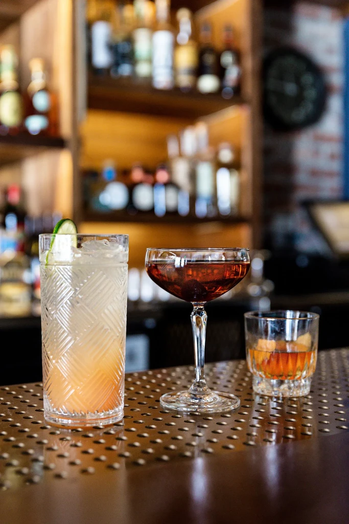 Three cocktails sit on a bar, bottles blurred in background.