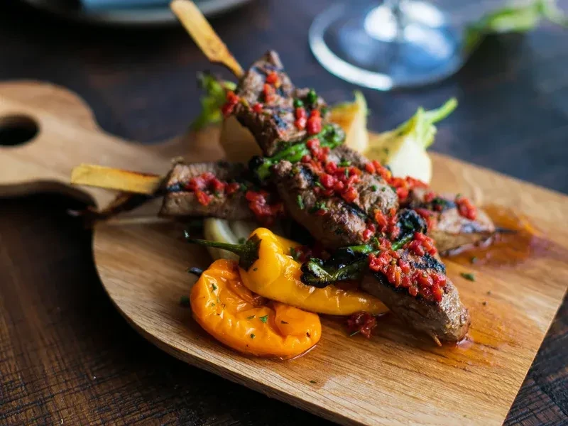 Grilled meat skewers with red salsa and yellow peppers on a wooden board.