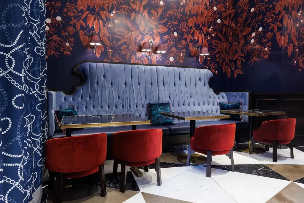 A restaurant with a blue velvet booth, red chairs, and botanical wallpaper.