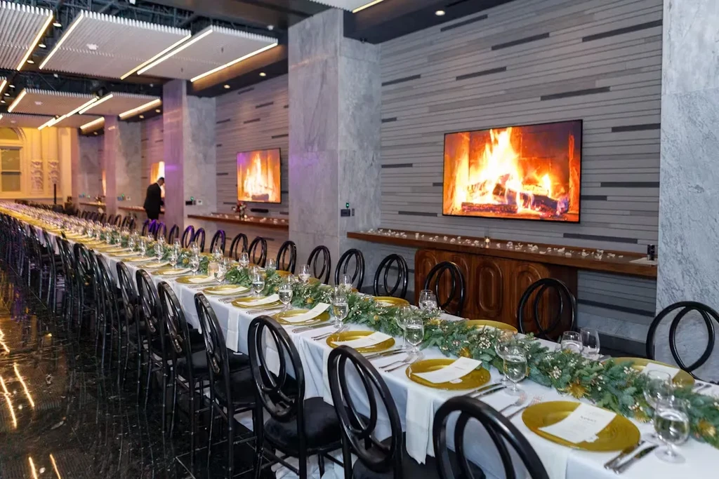 Long dining table with place settings, garlands, and two large TV screens.