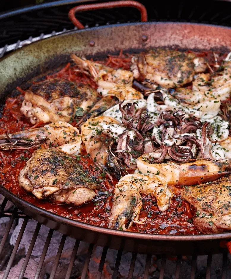 A large paella pan filled with chicken, shrimp, and squid on a grill.