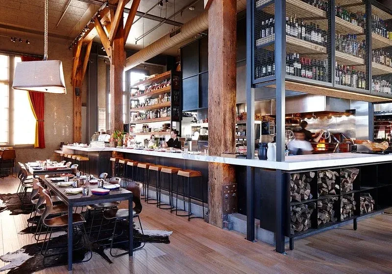 Rustic industrial restaurant with open kitchen and stacked firewood.
