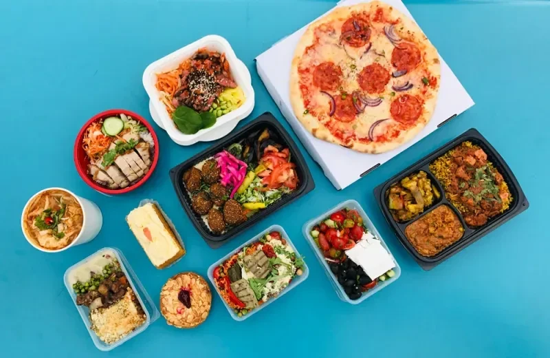Various takeout food containers, including pizza, salad, and main courses, on a blue surface.