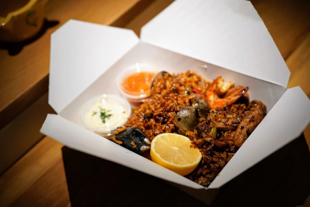 Seafood paella in a white takeout container with sauces and lemon.