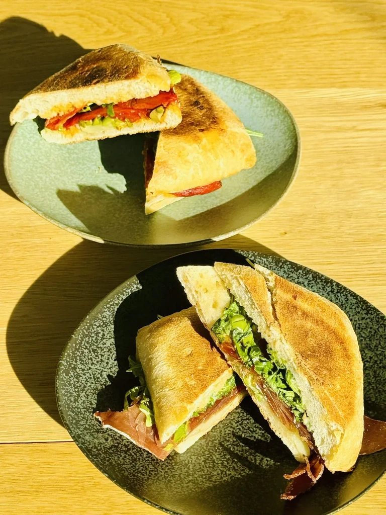 Two plates of Italian-style sandwiches on a light wood surface.