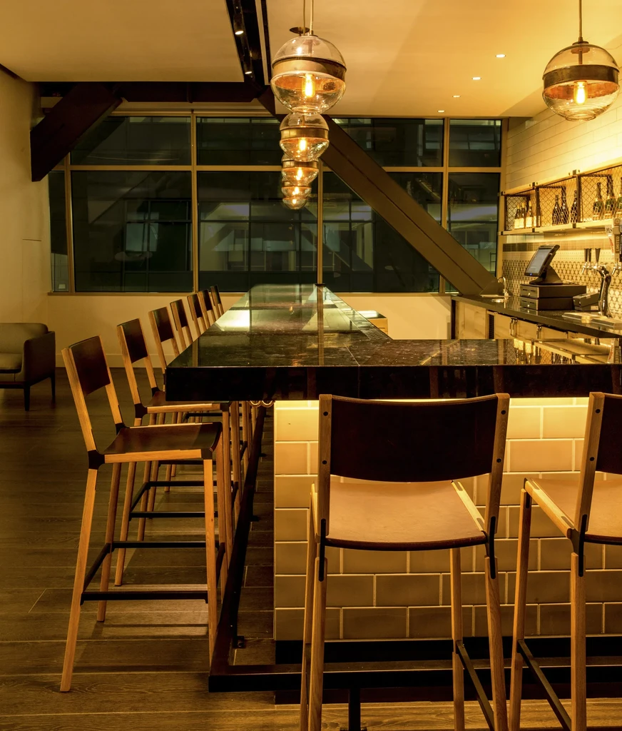 Modern bar with a long dark countertop, tall stools, and large windows.
