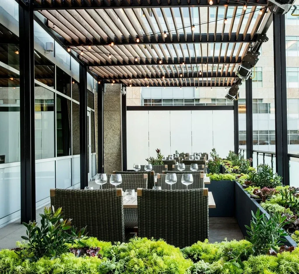 An outdoor restaurant patio with tables, chairs, plants, and a slatted roof.