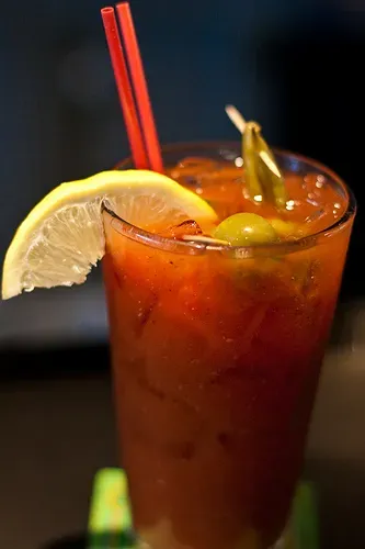 Tall glass of reddish drink with lemon, olives, green pepper, and straws.