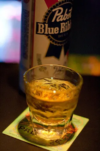 Glass of amber liquid with ice on coaster, Pabst Blue Ribbon can.