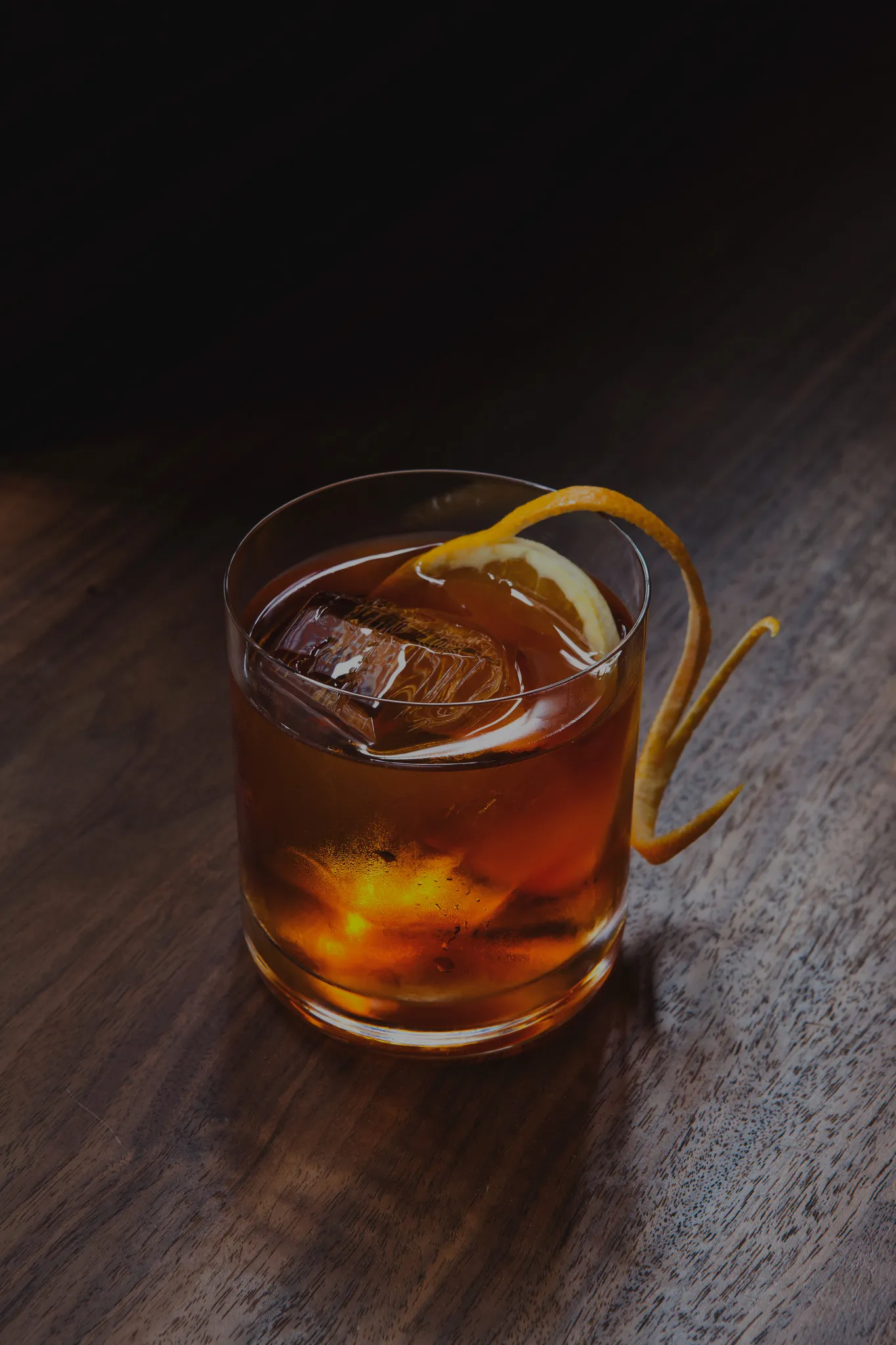 Amber cocktail in a glass with large ice and orange twist.