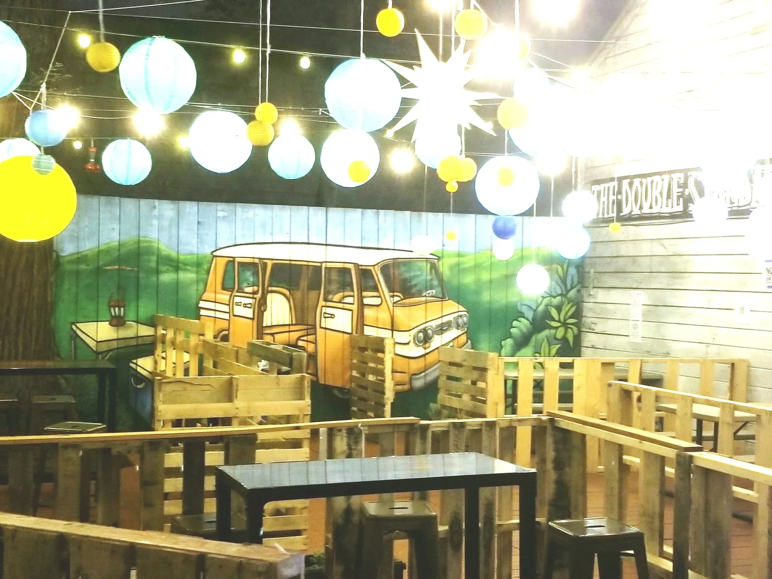 Colorful lanterns hang over a mural of a van and wooden pallet seating.