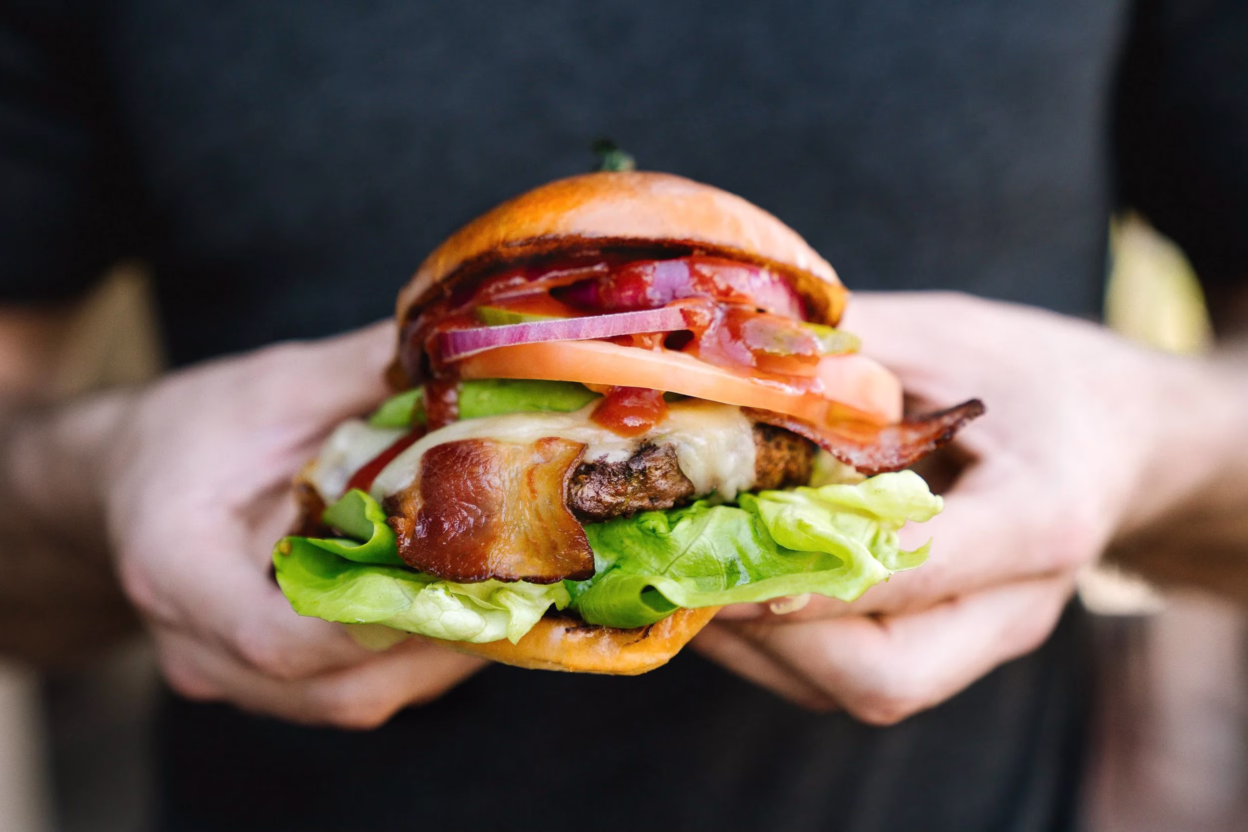 Hands holding a burger with bacon, cheese, lettuce, tomato, and onion.