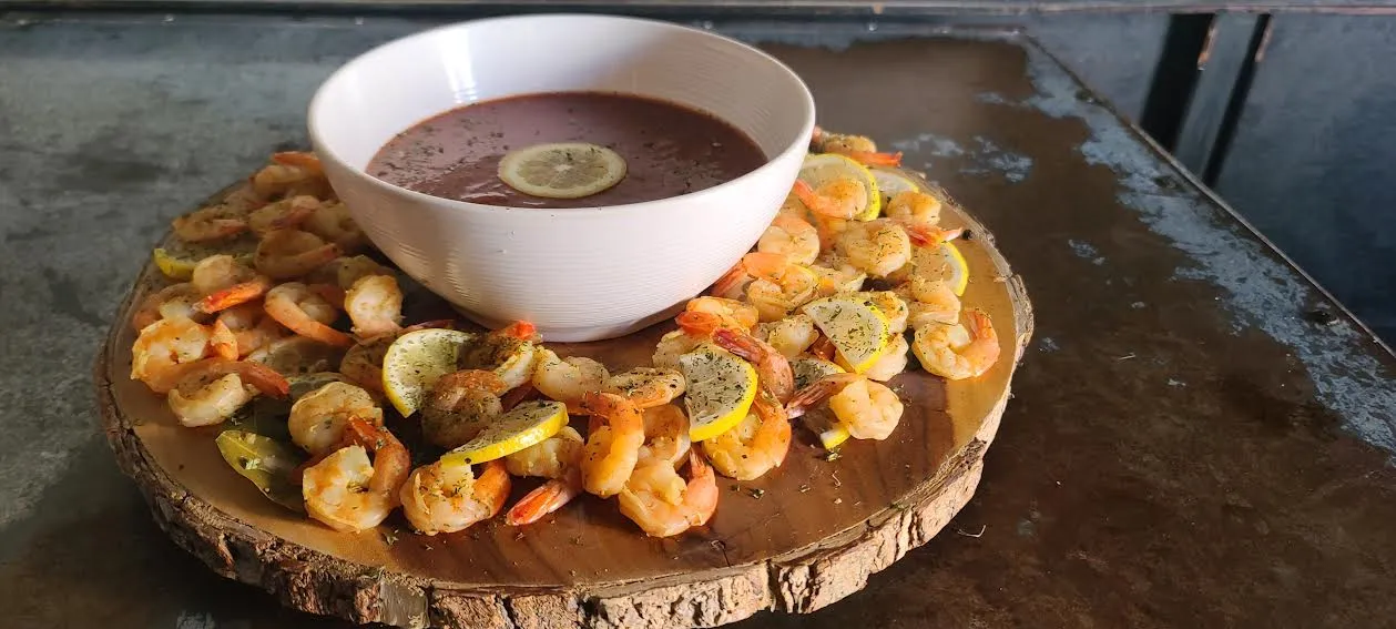 Seasoned shrimp and lemon surrounding a bowl of sauce on wood.