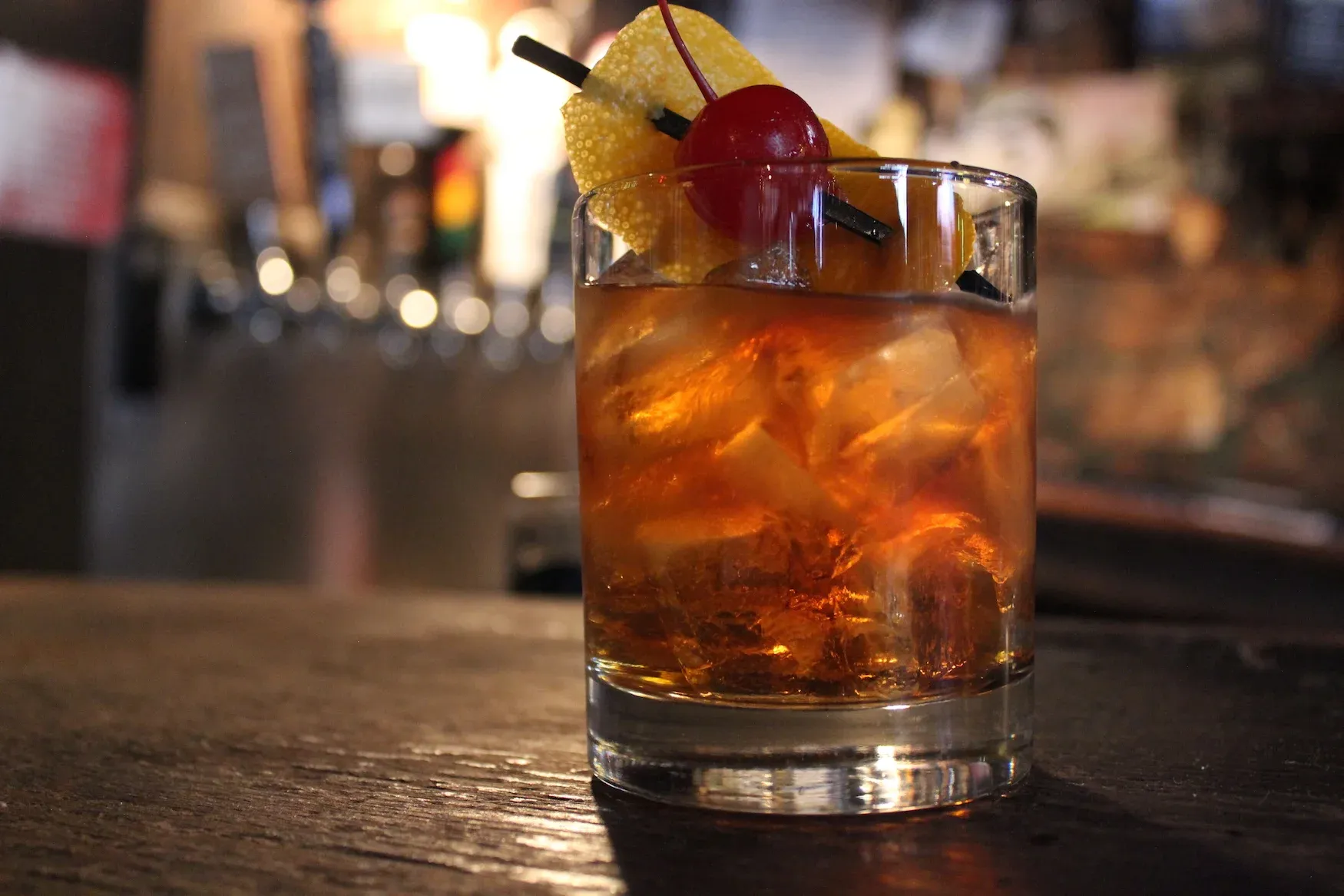 Glass of iced amber drink with orange peel and cherry.