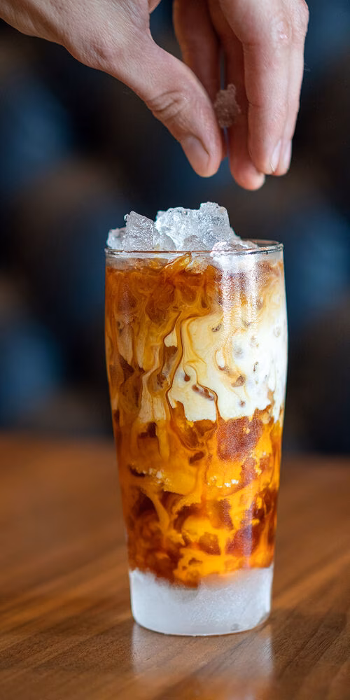 A hand adds ice to a swirled amber and cream iced beverage.