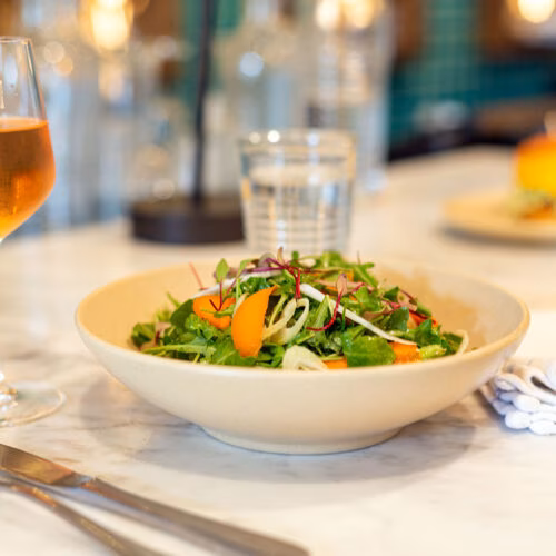 A bowl of mixed green salad with orange slices on a marble table.