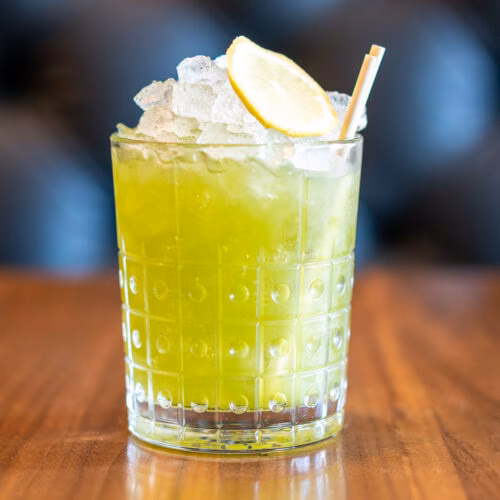 Green drink in a textured glass with ice, lemon, and straw.