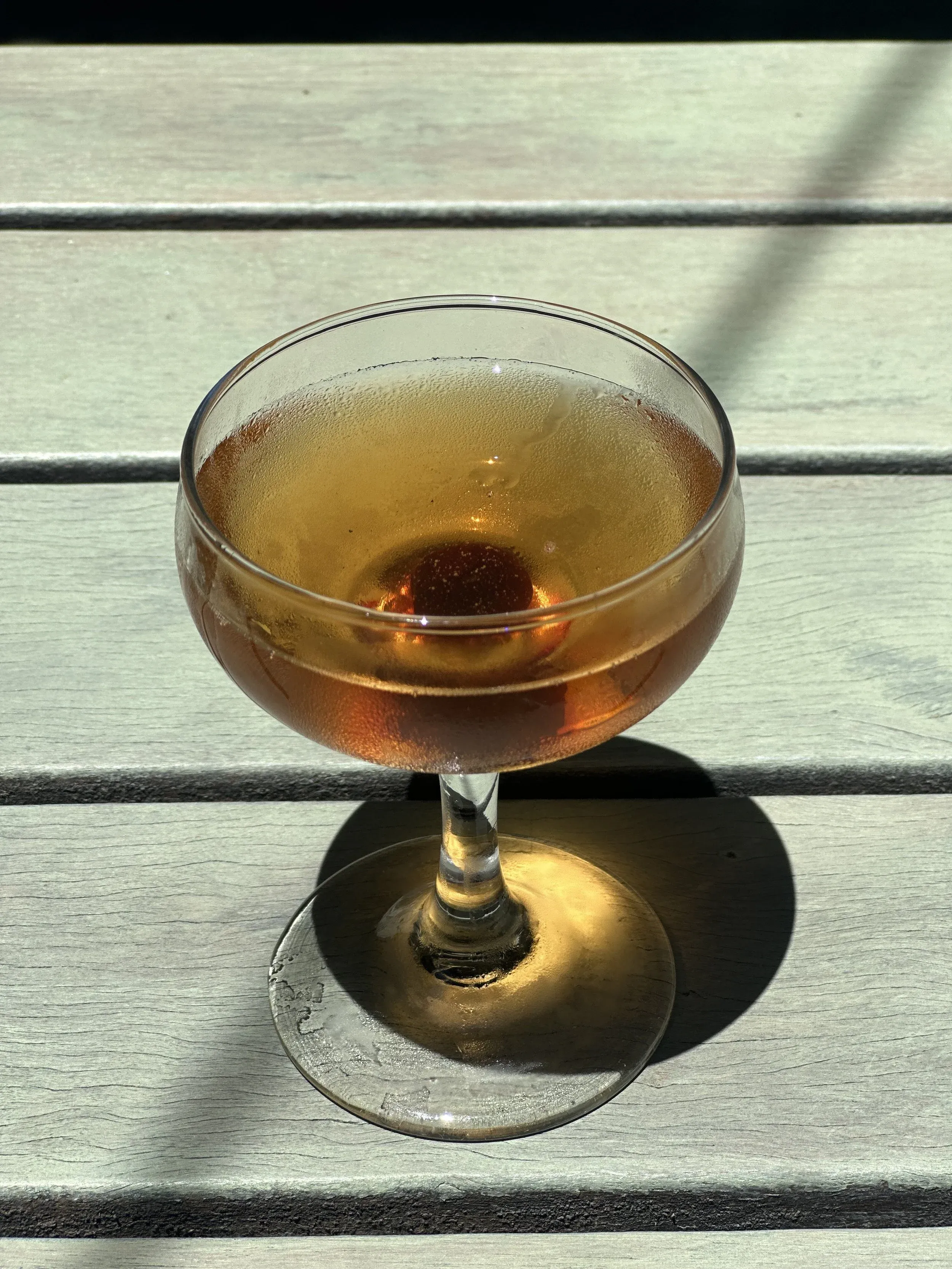 Frosted coupe glass with amber drink and cherry on a wooden table.