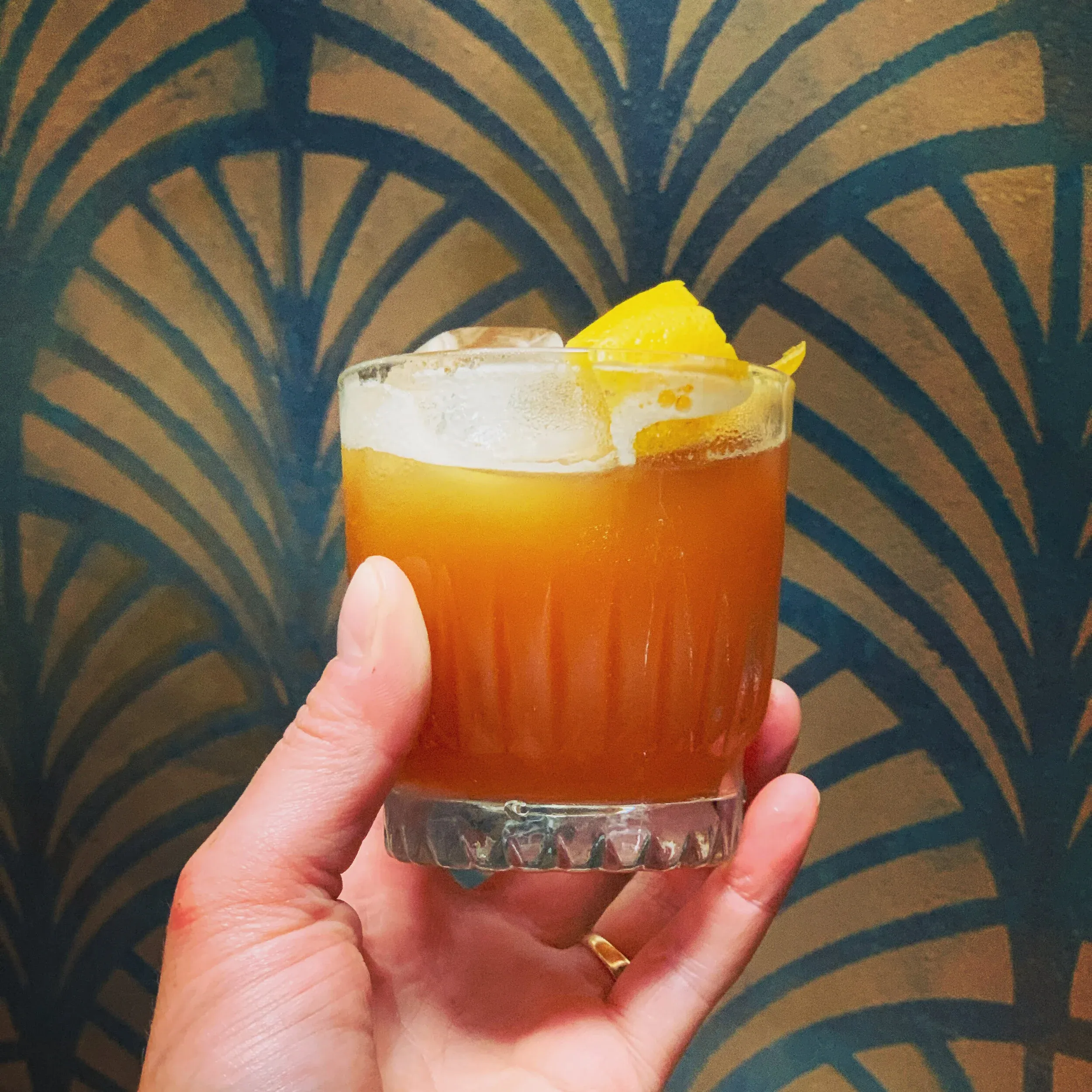 Hand holds an orange cocktail with ice, lemon peel, and foam.
