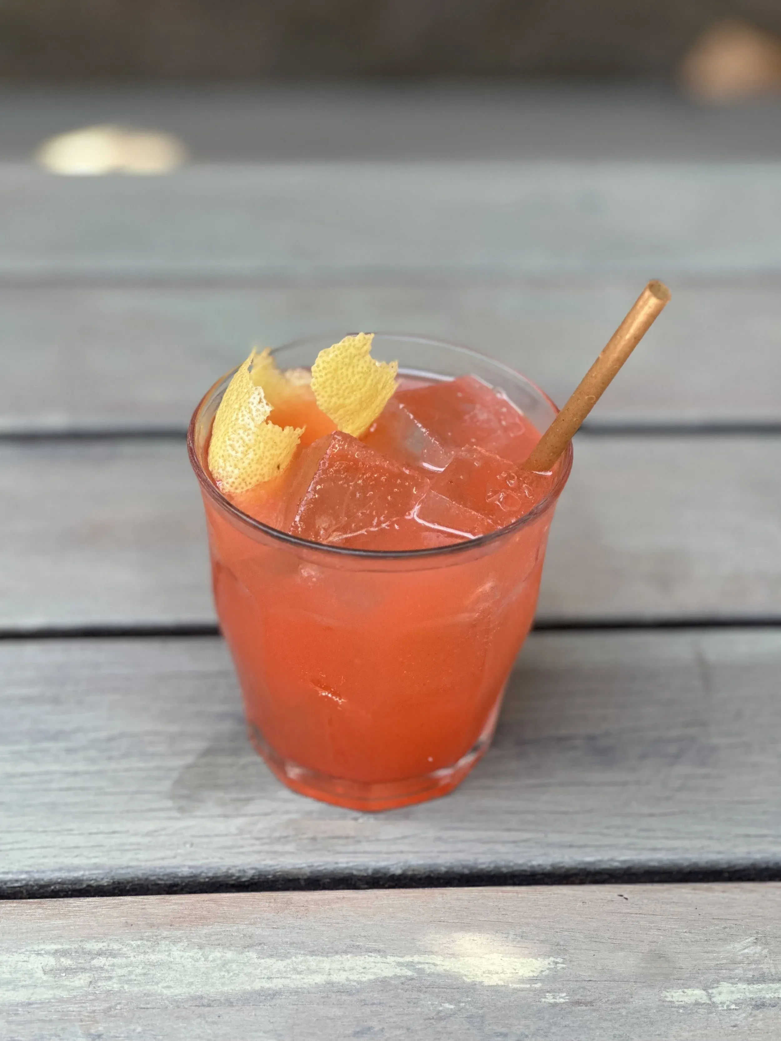 Glass with reddish-orange drink, ice, lemon peel, and straw.